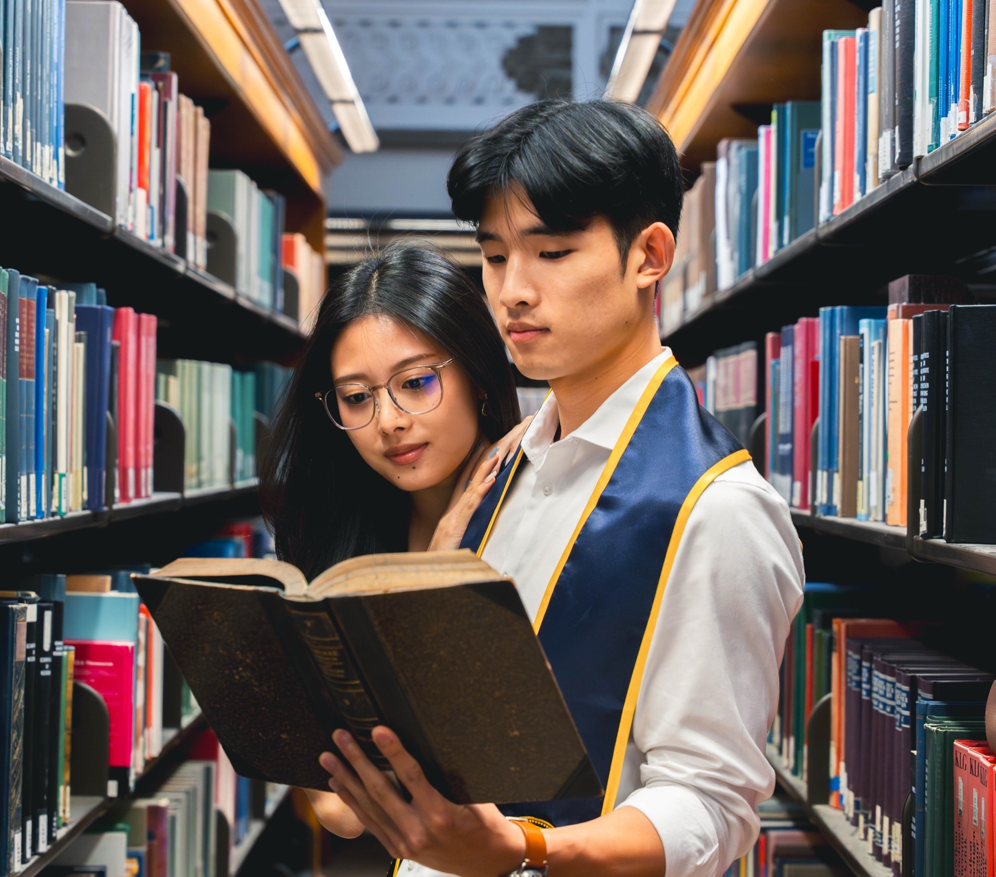 Two graduates reading an old book together in the library stacks