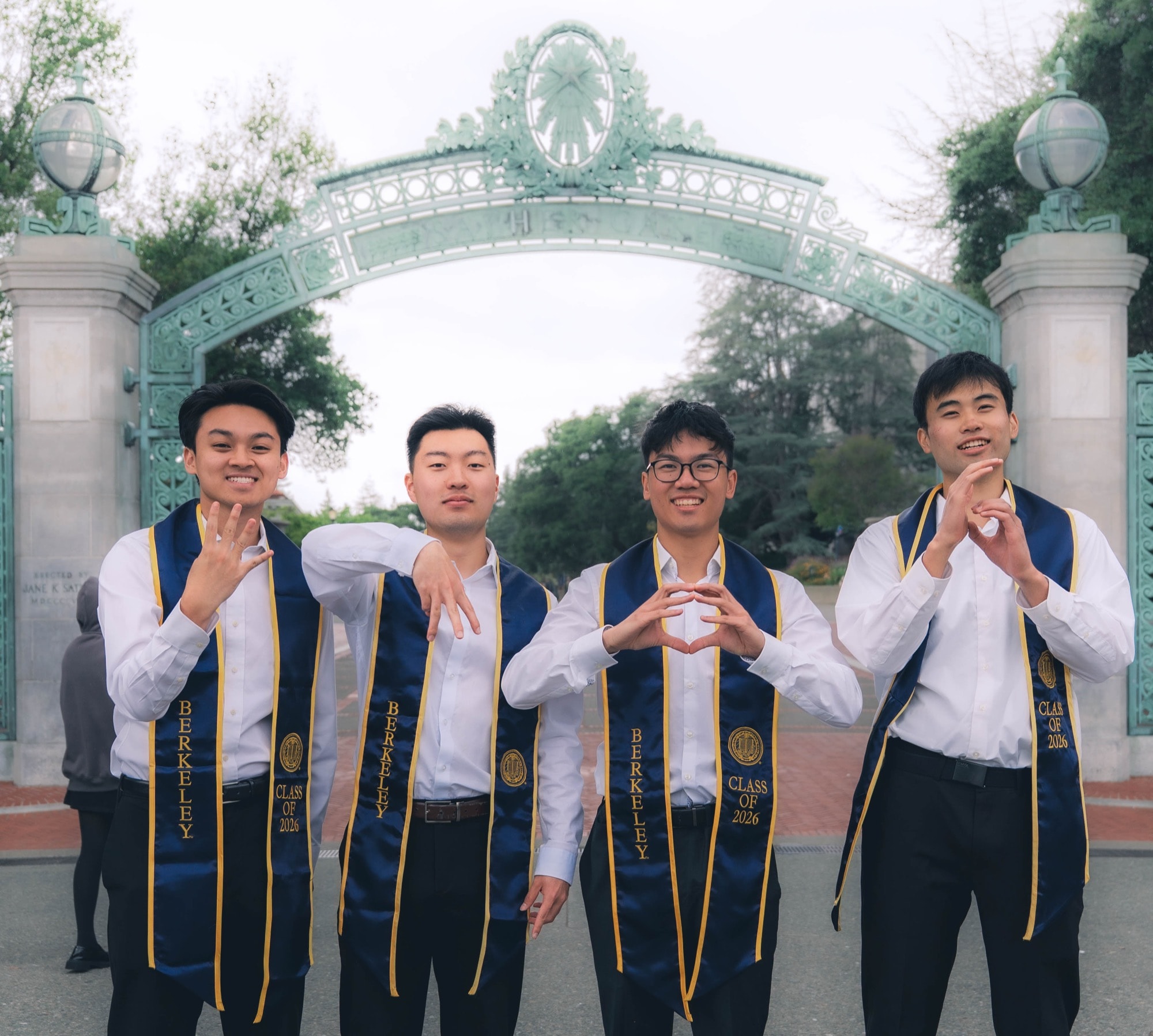 Four graduates throwing hand signs beneath Sather Gate