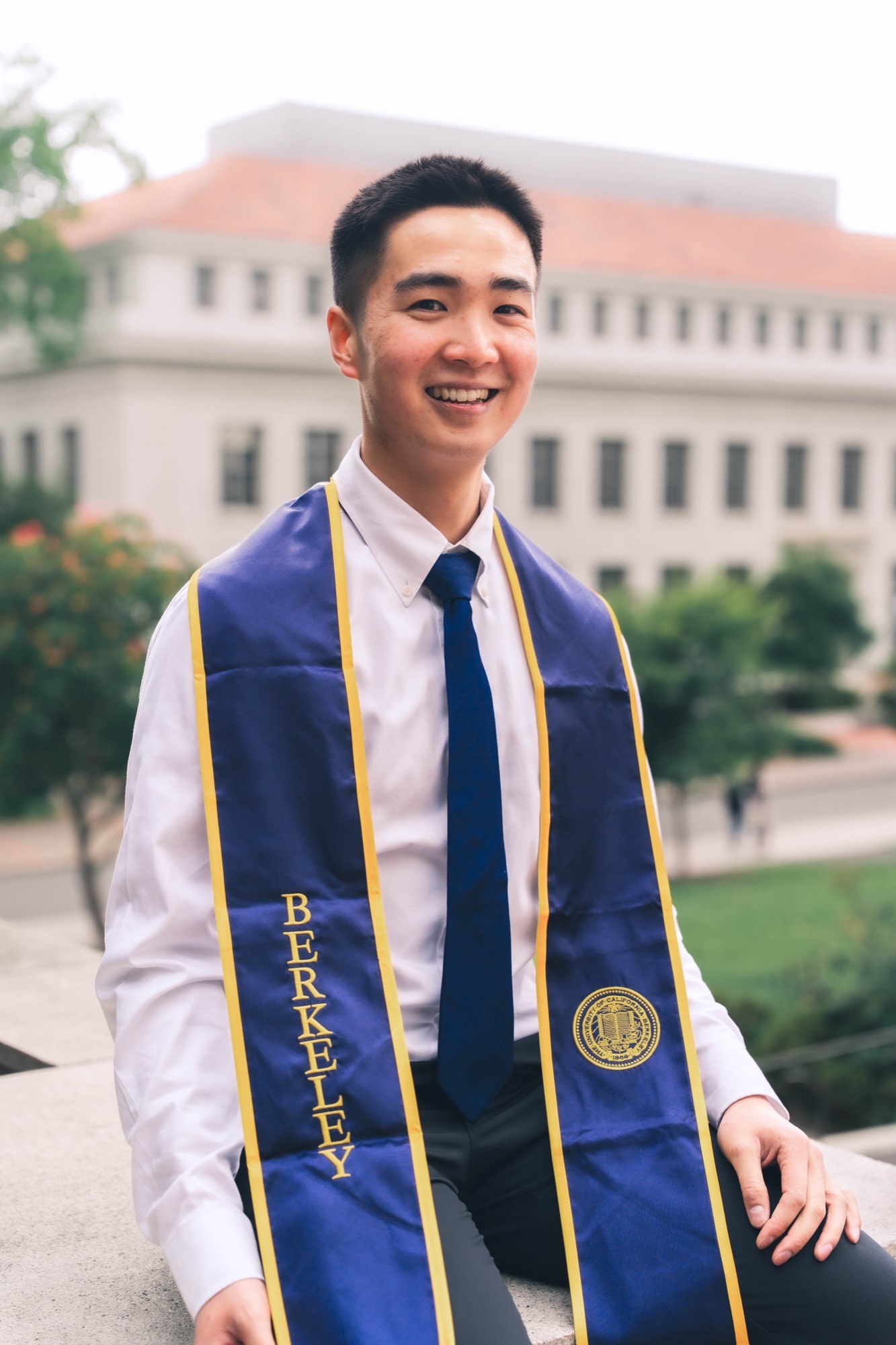Graduate smiling on a ledge, campus building soft behind in daylight